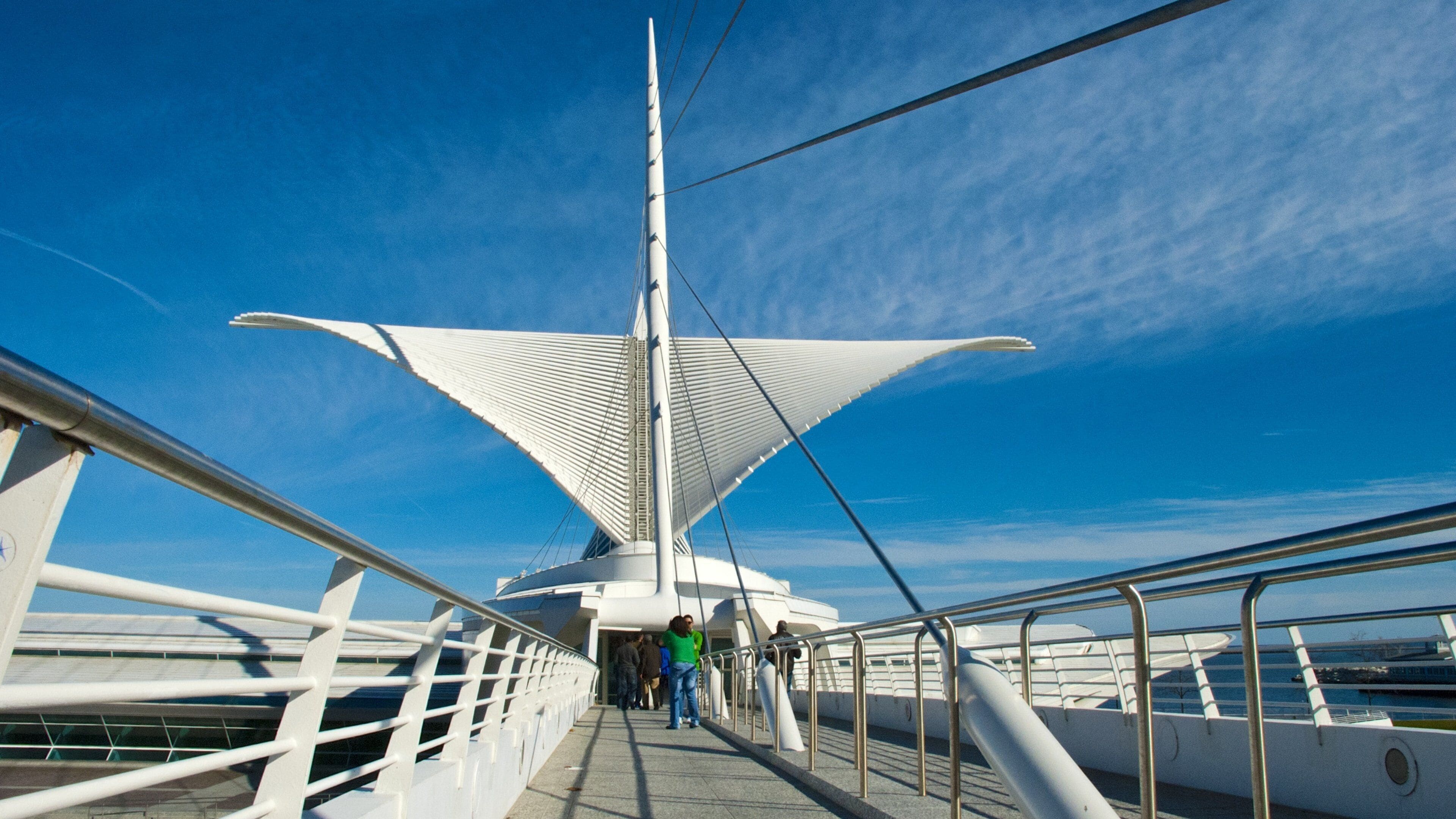 A pedestrian bridge with a distinctive white sail-like structure against a clear blue sky.
