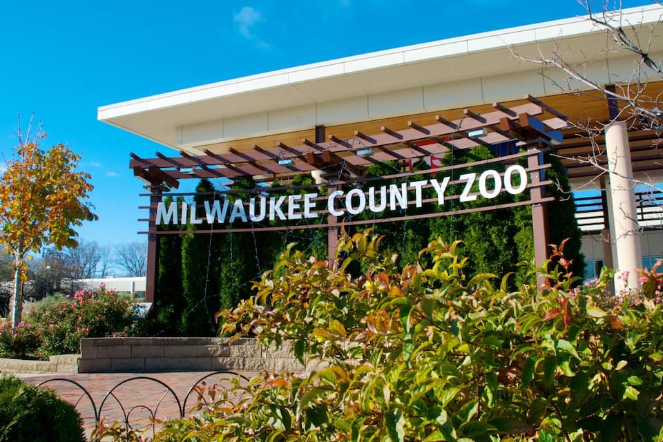 Entrada do Zoológico do Condado de Milwaukee, com uma placa, rodeada por árvores e plantas.