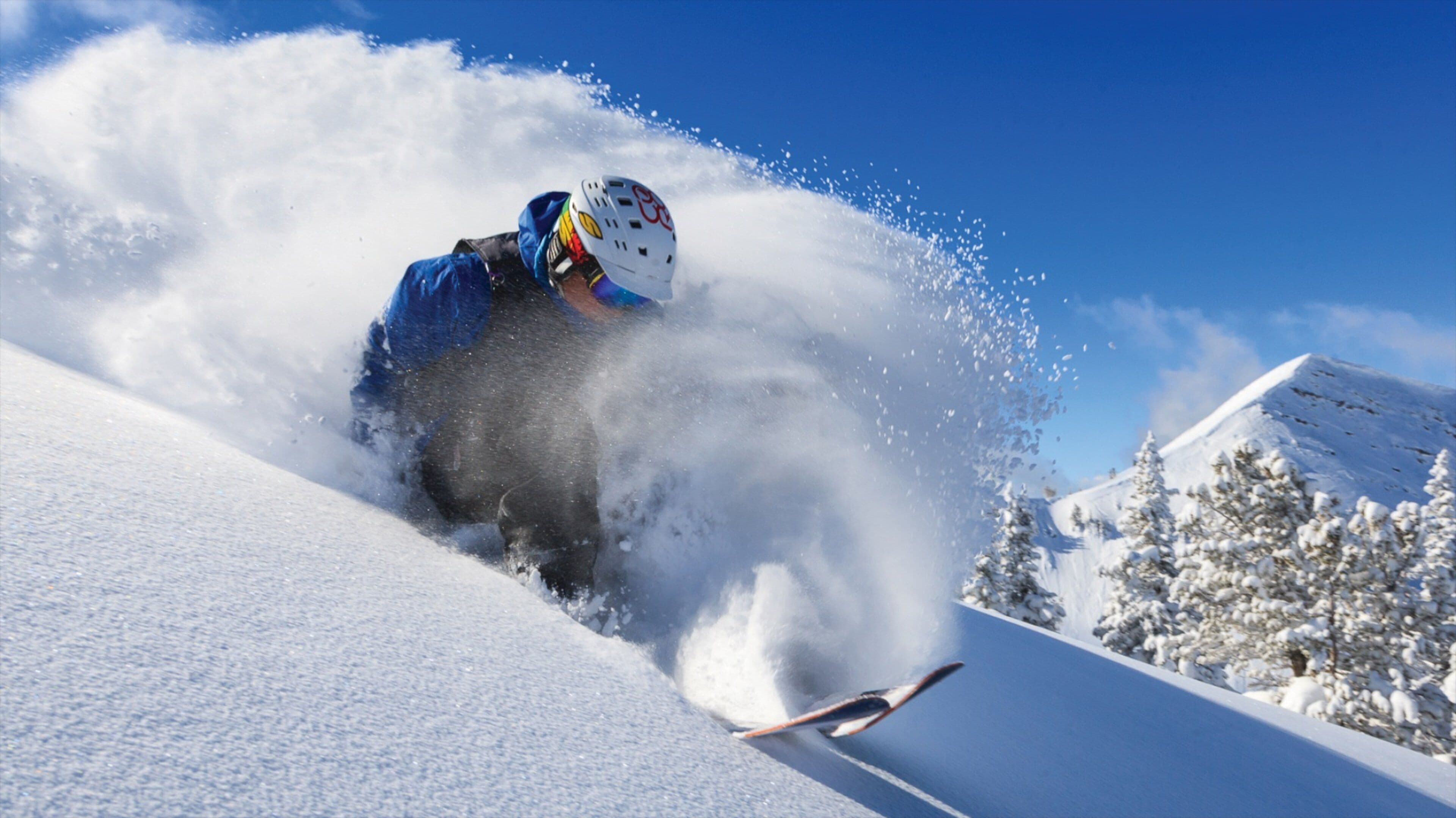 A skier in a blue jacket and helmet, carving a turn on a snowy slope with trees in the background.
