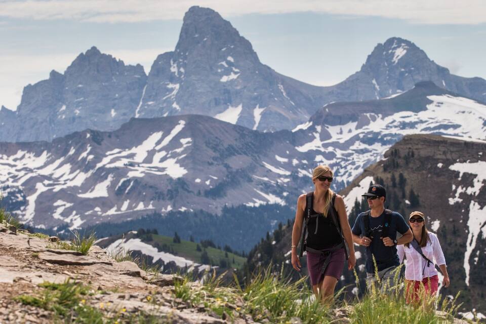 A group of hikers on a rocky mountain trail with snow-capped peaks in the background.