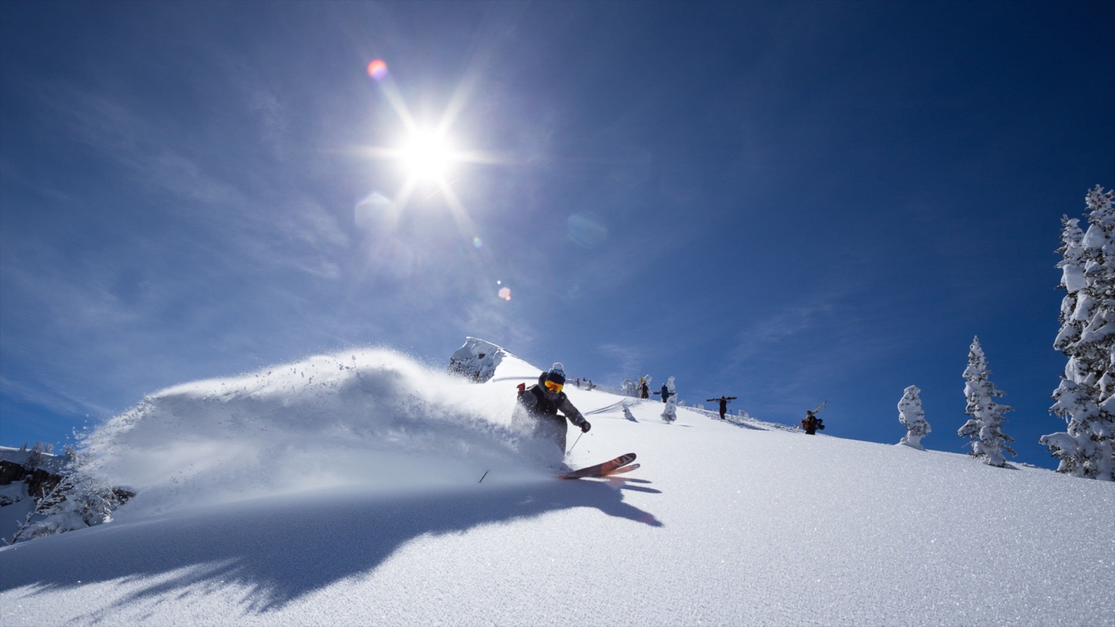 A skier descending a snow-covered slope with a clear blue sky and sun in the background.