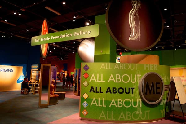 An exhibition with displays on origins and a large sign reading "ALL ABOUT HER ALL ABOUT ME".