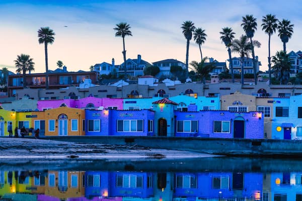 A coastal town with colorful buildings, palm trees, and a calm body of water reflecting the architecture.