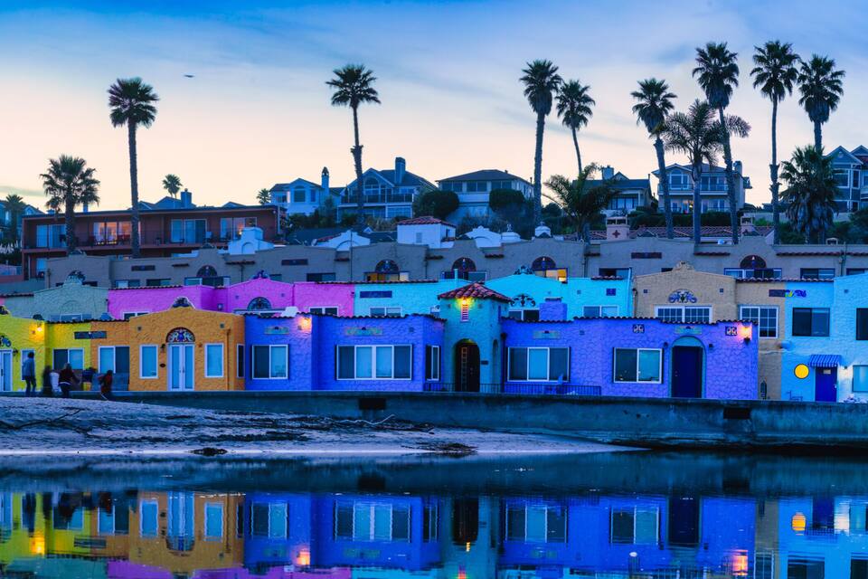 A coastal town with colorful buildings, palm trees, and a calm body of water reflecting the architecture.