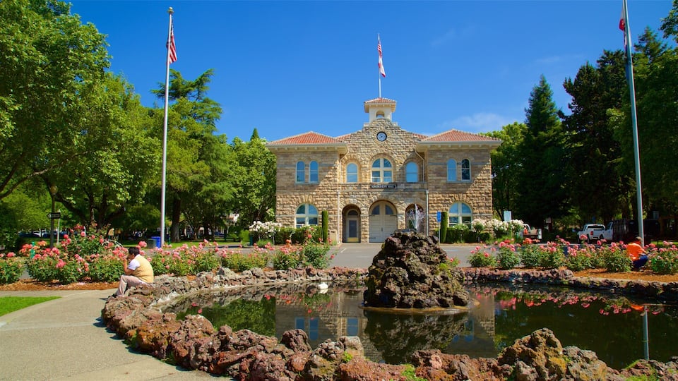 A stone building with arched windows and a clock tower, surrounded by a garden with a pond and flowers.