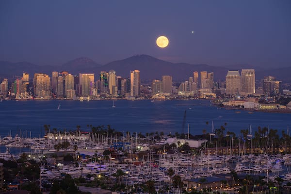 A city skyline with a prominent full moon in the sky, a marina with numerous boats, and a mountain range in the background.