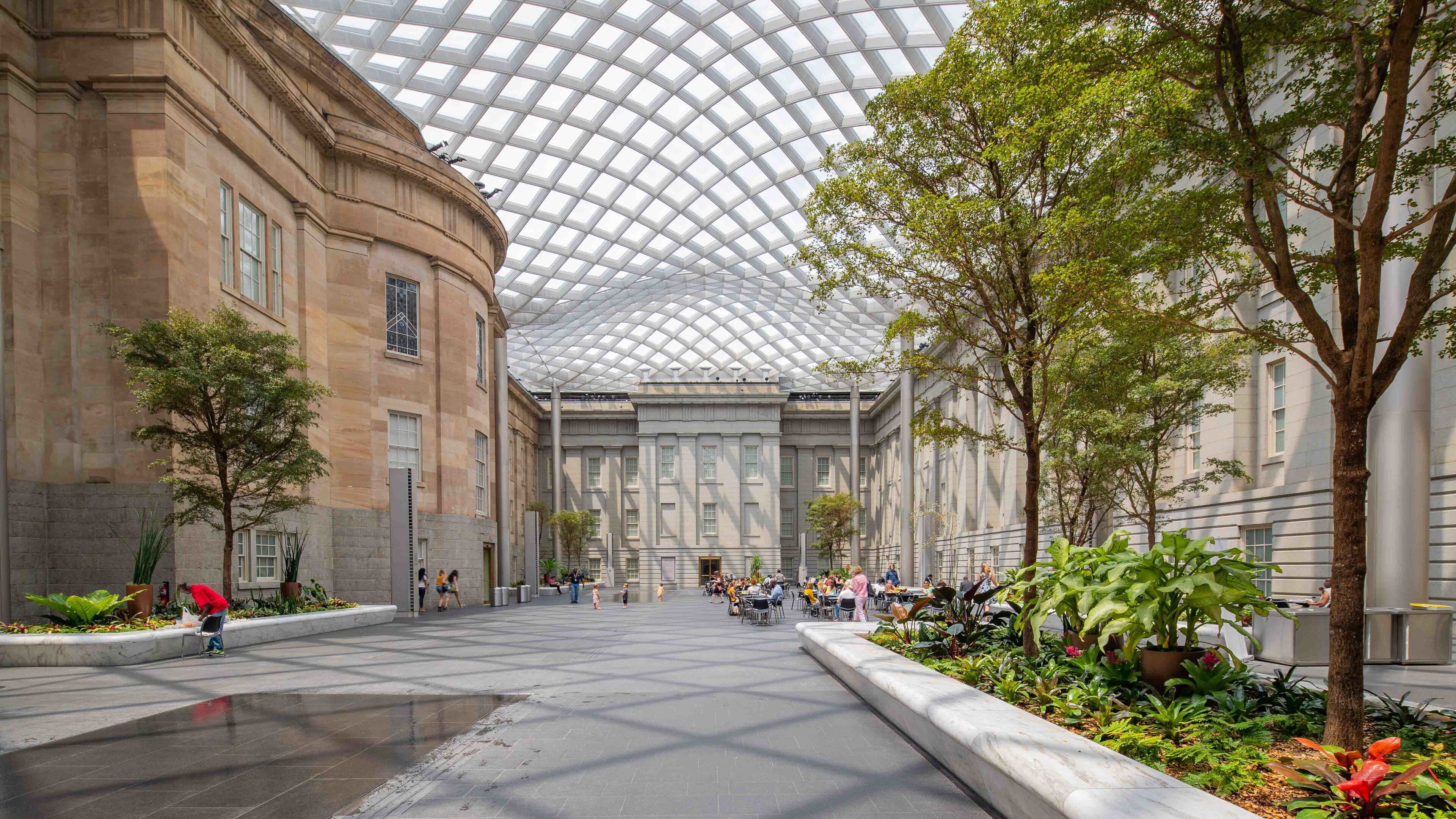 A spacious indoor courtyard with a glass dome ceiling, surrounded by classical architecture and lush greenery.