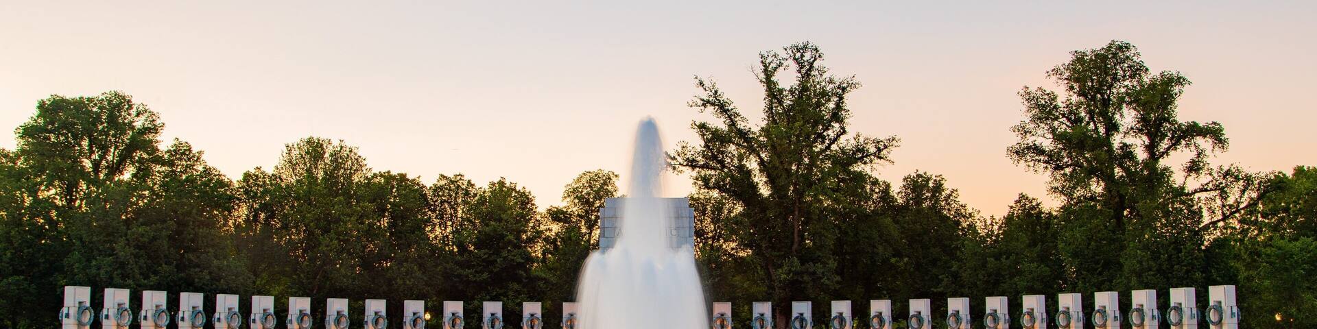 A large fountain with water jets in a circular pattern, surrounded by trees and a row of memorial columns.