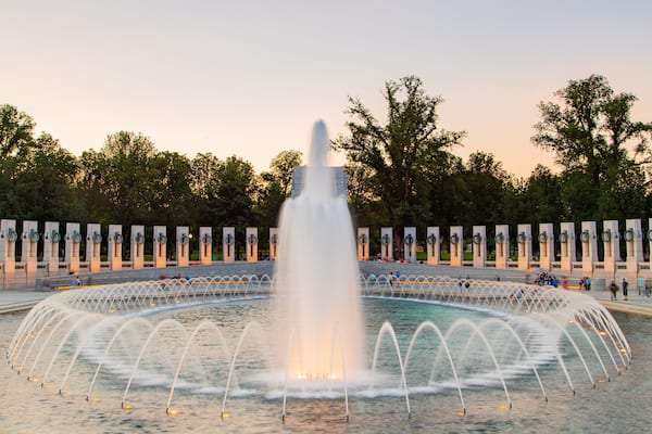 Une grande fontaine avec des jets d’eau disposés en cercle, entourée d’arbres et d’une rangée de colonnes commémoratives.