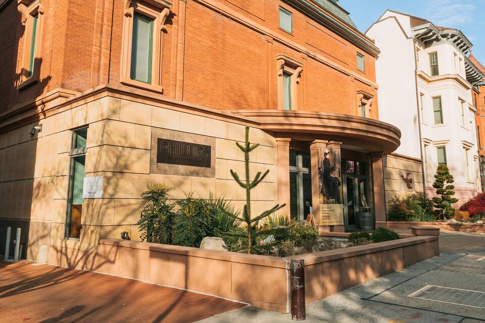 A classical building with a plaque reading "PHILLIPS The Phillips Collection" and a small tree in the planter.