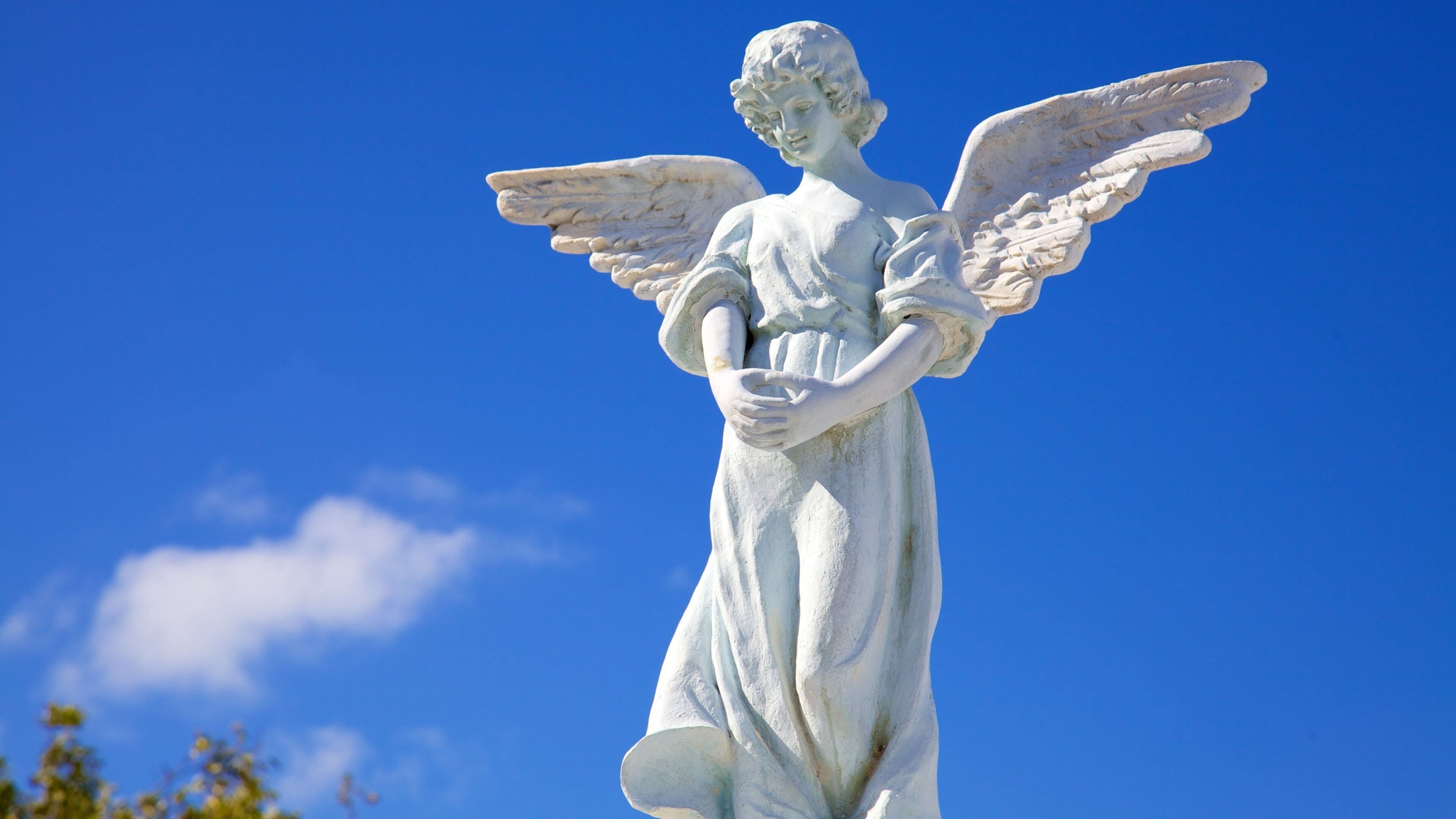 Una statua bianca di un angelo con le ali, che tiene un libro, sullo sfondo di un cielo azzurro.