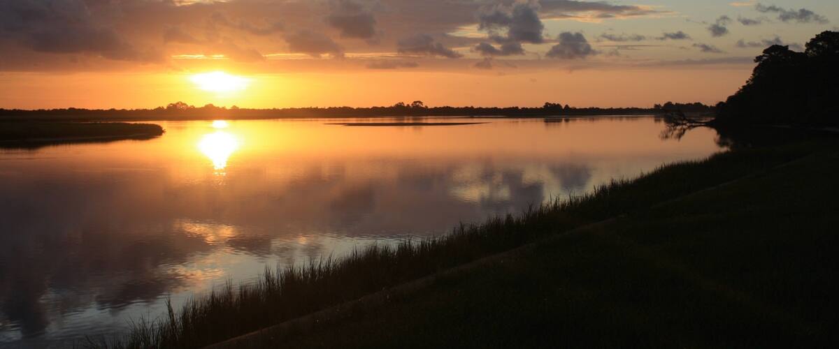A sunset over a calm river with a silhouette of trees and a distant landmass.