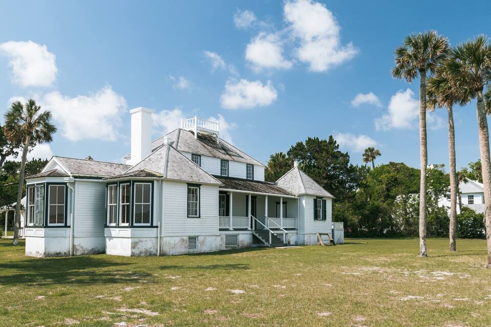 Una casa blanca de estilo colonial con un porche y chimenea, rodeada de palmeras y césped.