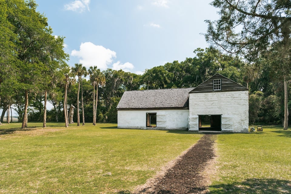 A white building with a dark roof, surrounded by trees and a grassy field.