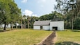 Un edificio blanco con techo oscuro, rodeado de árboles y un campo cubierto de hierba.