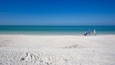 A sandy beach with clear blue water and a few people walking.