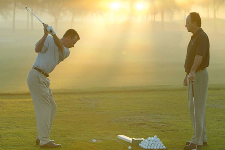 A golf course with a player preparing to hit a ball, another observing, and a pile of golf balls on the ground.