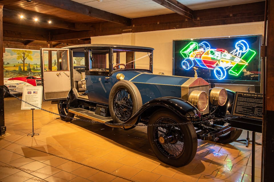 A vintage car on display in a museum with a neon sign in the background.
