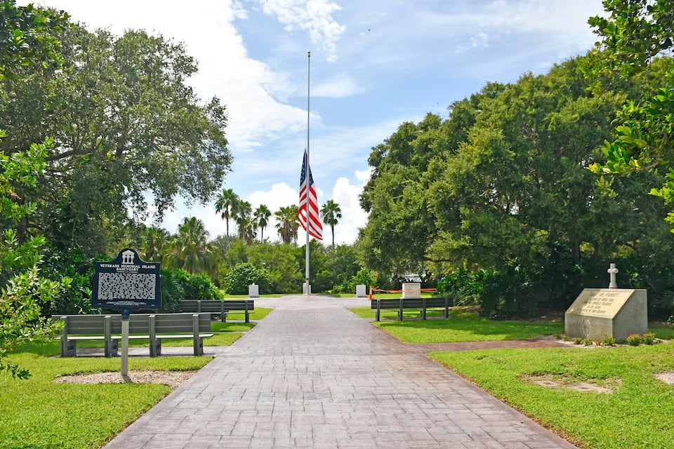 A Veterans Memorial Island Sanctuary pathway with an American flag, benches, and a signboard.