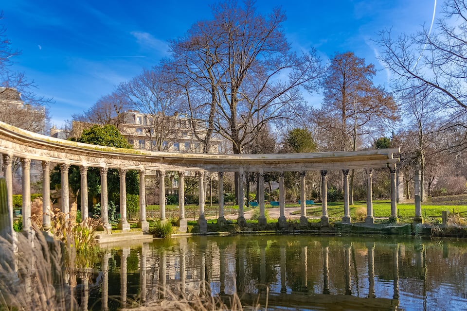 Uma ponte clássica com colunas sobre um lago refletor, cercada por árvores e edifícios.