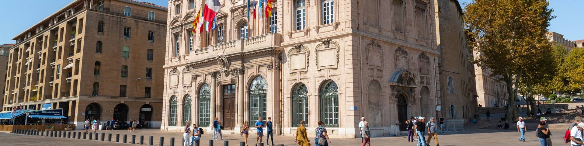 A historic building with multiple flags, large windows, and a paved plaza with bollards.