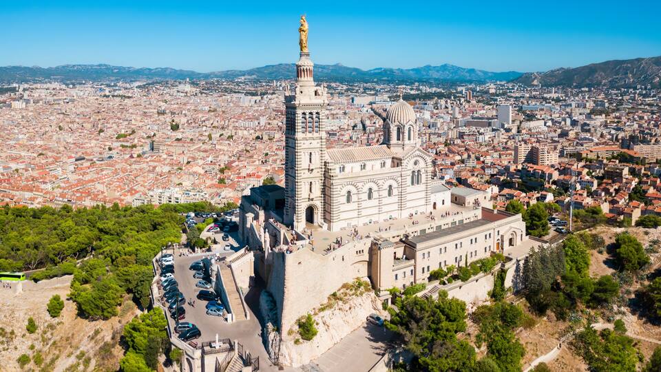 Una cattedrale con un'alta torre e una statua, immersa in un panorama urbano con montagne sullo sfondo.