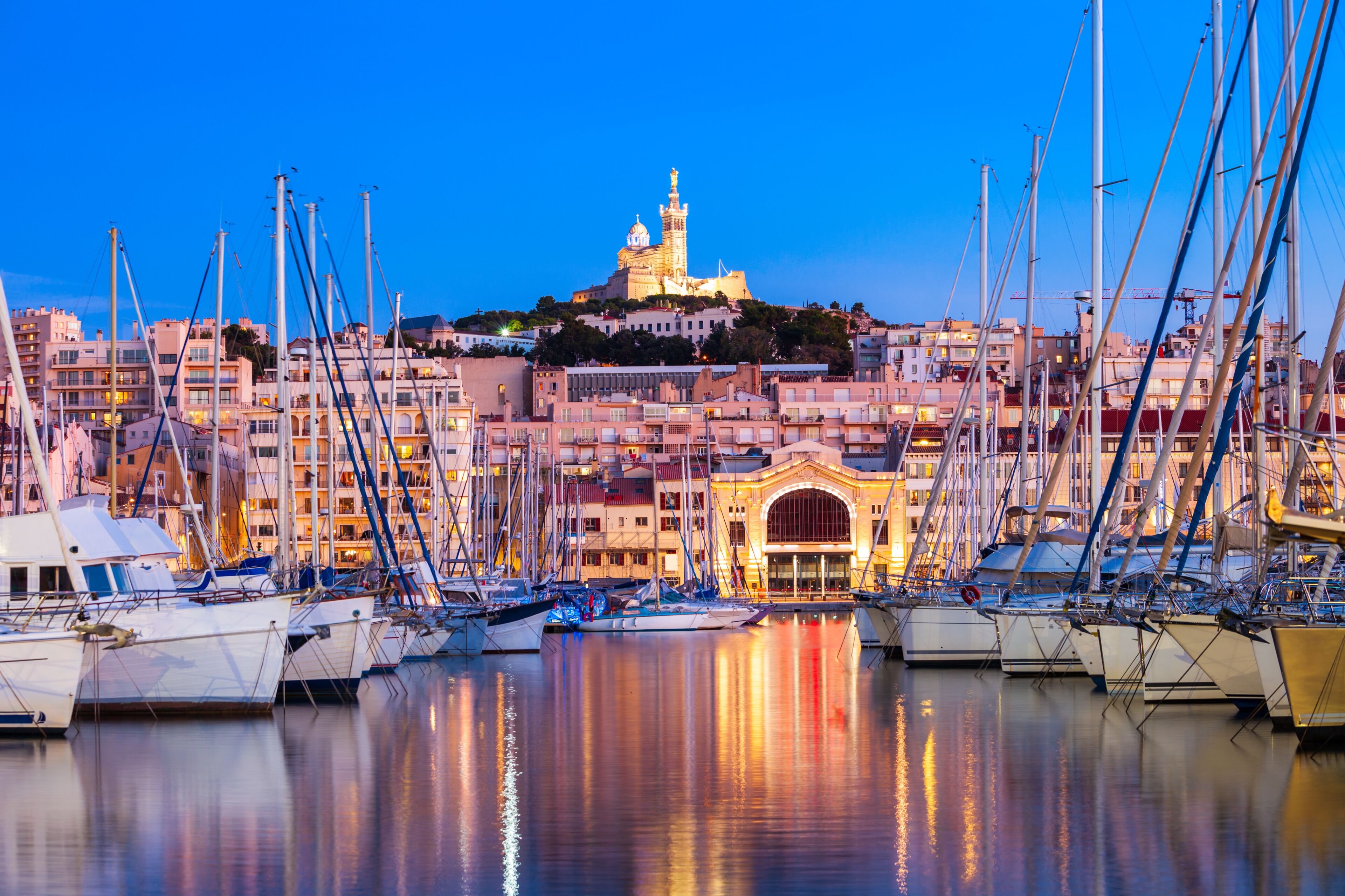 Una marina con numerosi velieri ormeggiati, un edificio importante con una cupola e una torre, e un panorama urbano sullo sfondo.