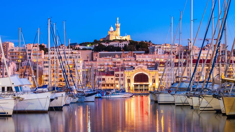 Una marina con numerosi velieri ormeggiati, un edificio importante con una cupola e una torre, e un panorama urbano sullo sfondo.