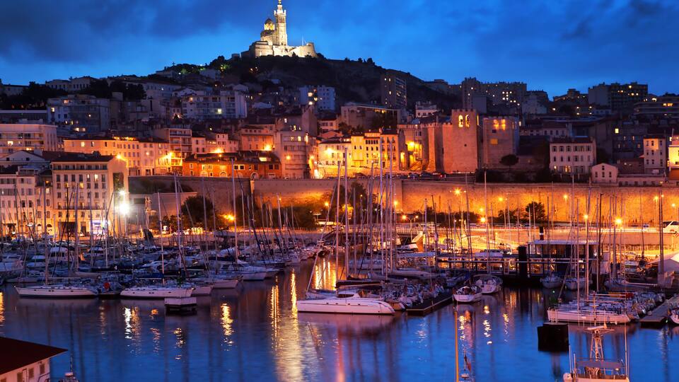 Una marina al crepuscolo con barche ormeggiate, una torre imponente su una collina e un panorama urbano con edifici illuminati.