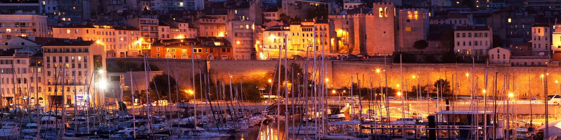 Un port de plaisance au crépuscule, avec des bateaux amarrés, une tour imposante sur une colline et un paysage urbain avec des bâtiments illuminés.