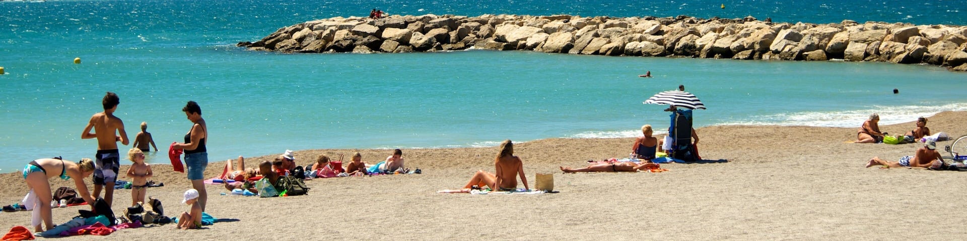 A sunny beach with people sunbathing and swimming, a rocky breakwater, and clear blue water.