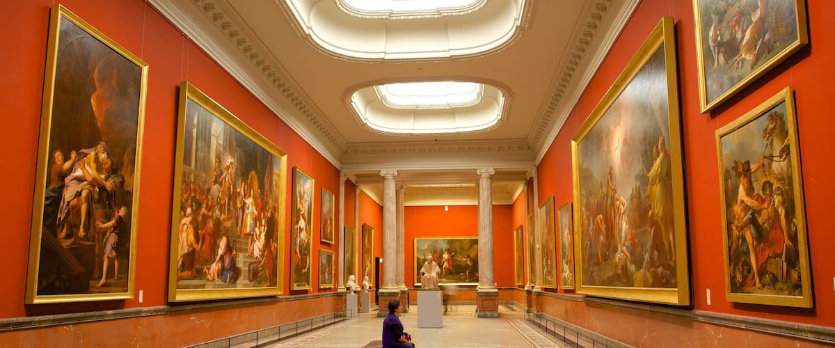 A museum corridor with large classical paintings on the walls and a marble floor.