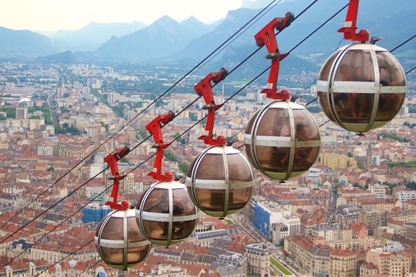 Un réseau de téléphériques avec des cabines sphériques surplombant une ville avec des bâtiments aux toits rouges et des montagnes en arrière-plan.