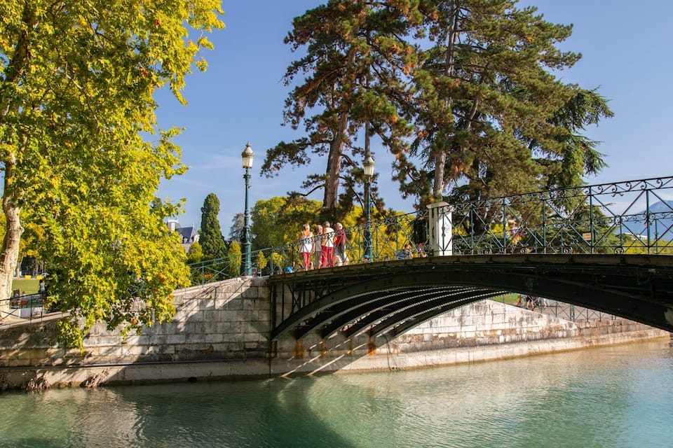A stone bridge over a calm river, surrounded by lush green trees and a clear blue sky.