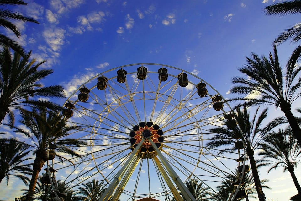 A large Ferris wheel with a star-shaped center, surrounded by palm trees under a blue sky with clouds.