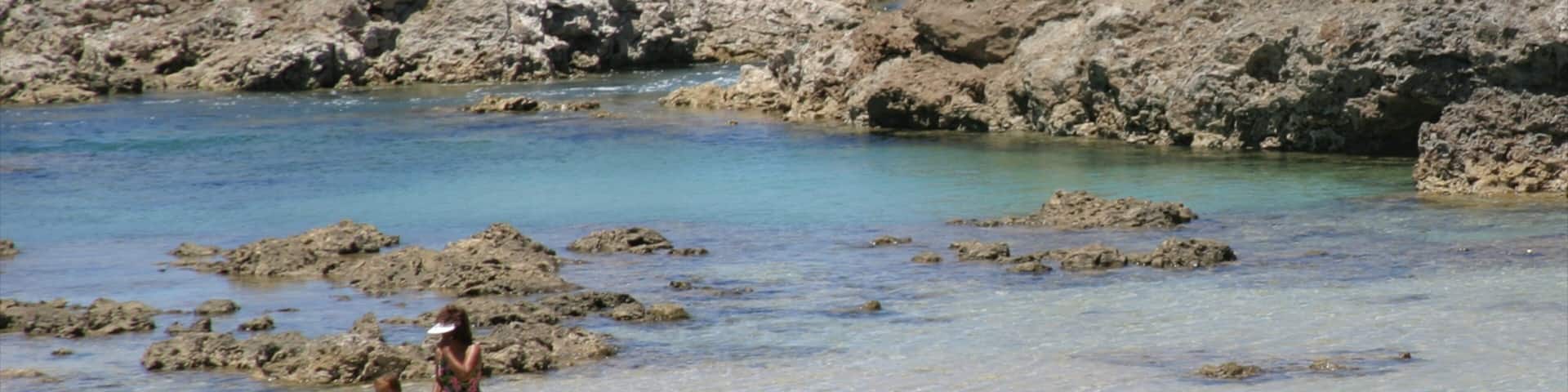 A rocky shoreline with clear water and a woman and child holding hands.