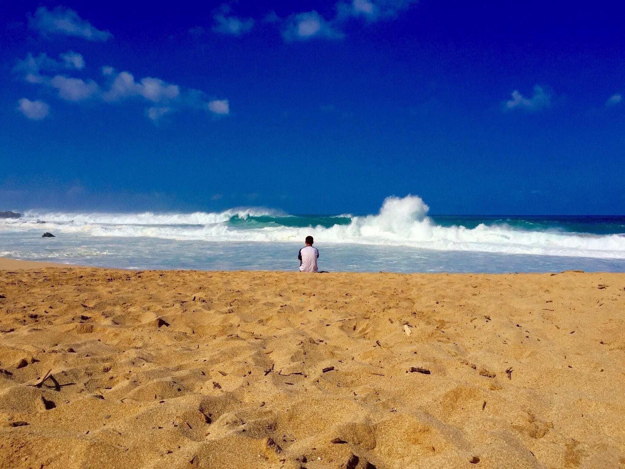 A person standing on a sandy beach facing the ocean with waves crashing onto the shore under a clear blue sky.