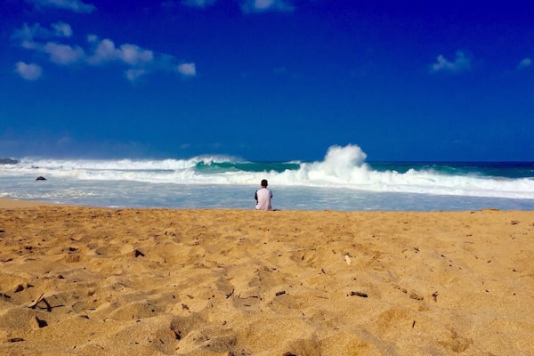 Uma pessoa está em pé na areia da praia, olhando para o oceano, com ondas quebrando na costa sob um céu azul claro.