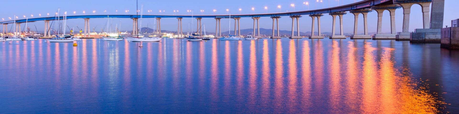 Un puente sobre aguas tranquilas con barcos y luces de la ciudad reflejándose en la superficie.