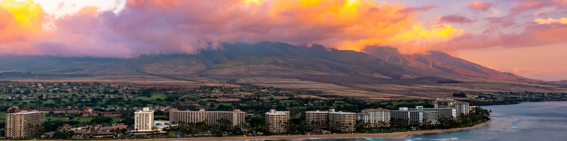 A coastal cityscape with a mountain range in the background, a marina with boats, and a beachfront hotel area.