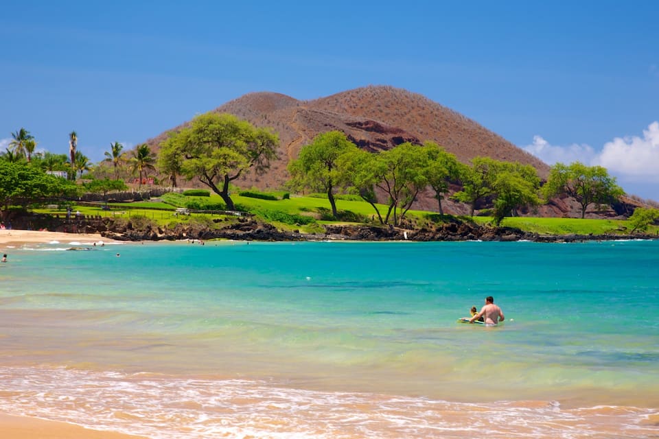A beach with clear turquoise water, a person in the water, and a mountain in the background.
