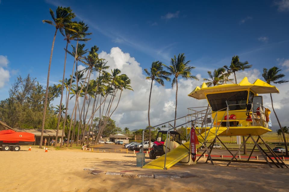 A lifeguard tower on a beach with palm trees and a red lifeguard boat in the background.