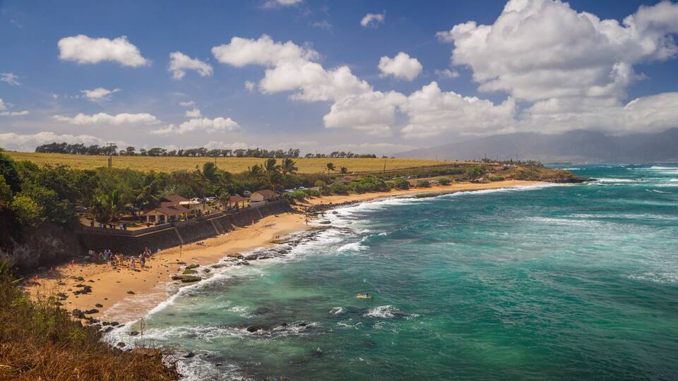 A coastal landscape with a sandy beach, turquoise ocean waves, and a clear sky with scattered clouds.