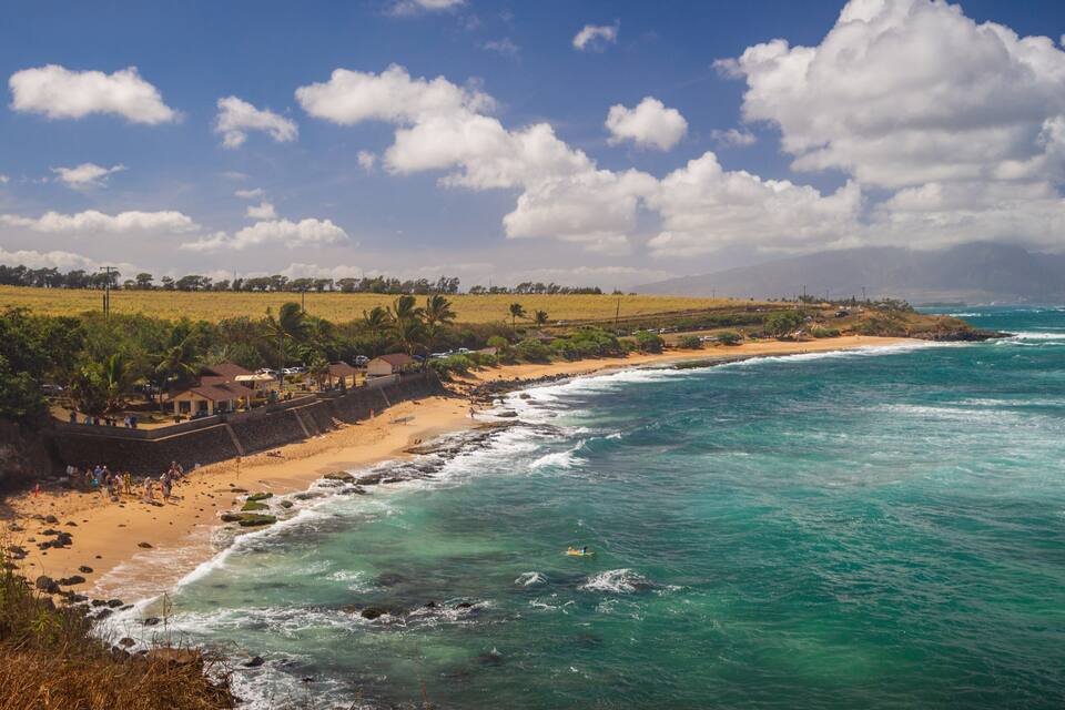 A coastal landscape with a sandy beach, turquoise ocean waves, and a clear sky with scattered clouds.