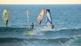 Tre windsurfere på et roligt hav under klare himmel.