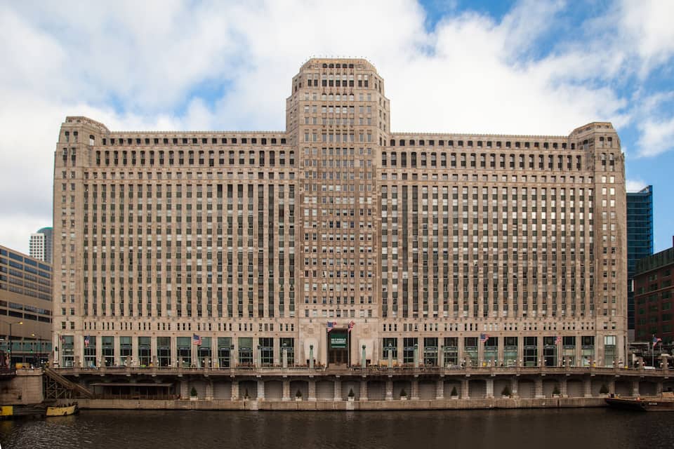 Un edificio grande y de estilo histórico con una torre central y numerosas ventanas, situado cerca de una masa de agua.