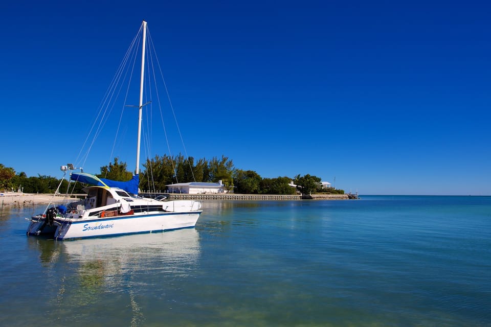 Un velero llamado 'Soundwave' fondeado en un puerto tranquilo con agua azul cristalina, rodeado de una playa de arena y vegetación exuberante.