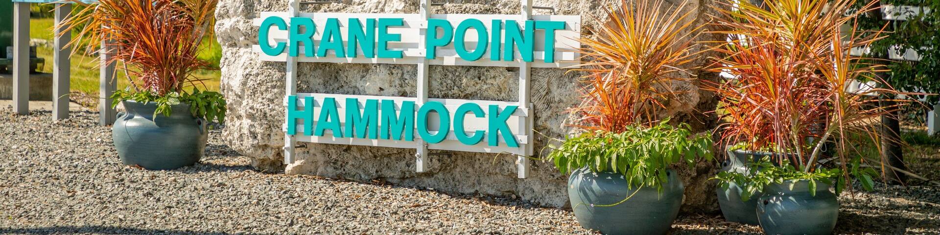 A sign for Crane Point Hammock with a tree logo and potted plants.