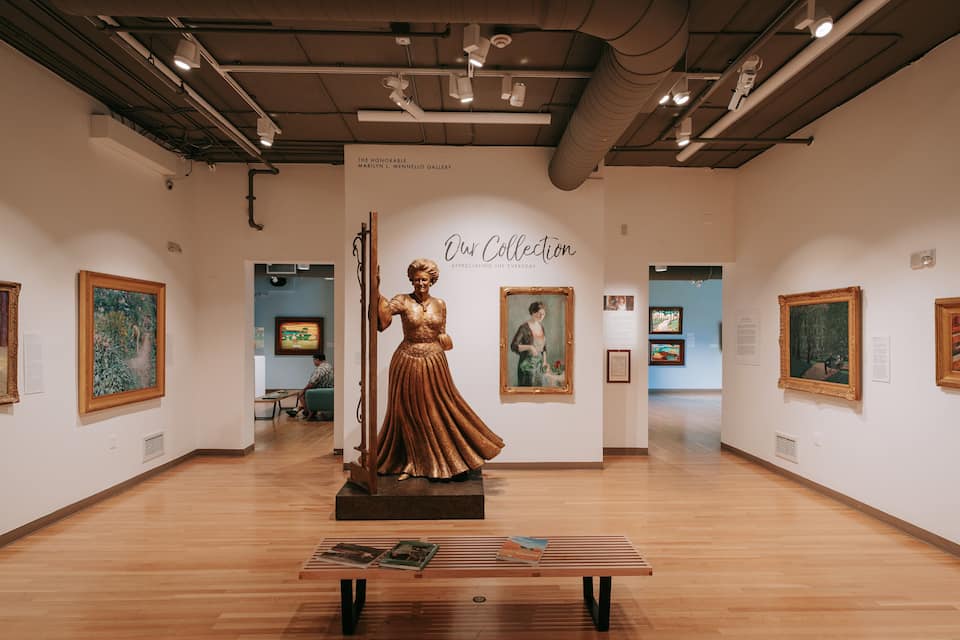 Een kunstgalerie met een centraal beeld, ingelijste schilderijen en informatieborden.