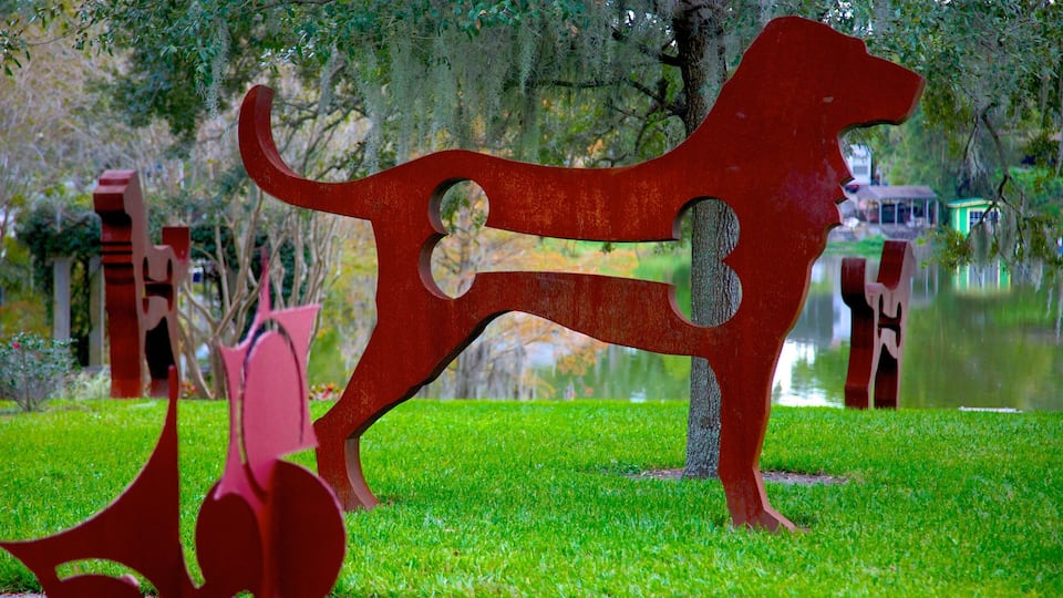 A red abstract sculpture of a dog in a park with a pond and trees in the background.